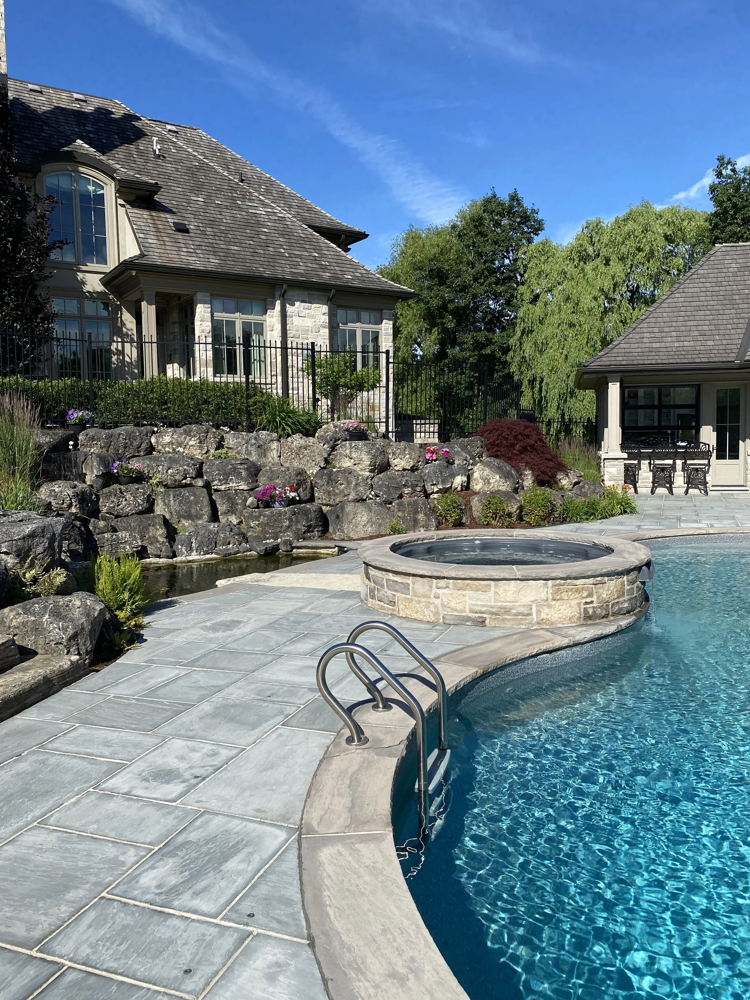 Backyard with swimming pool and hot tub, stone patio, rock wall, landscaped garden with flowers and shrubs, large house with stone and siding exterior, patio furniture, trees, and a clear blue sky.
