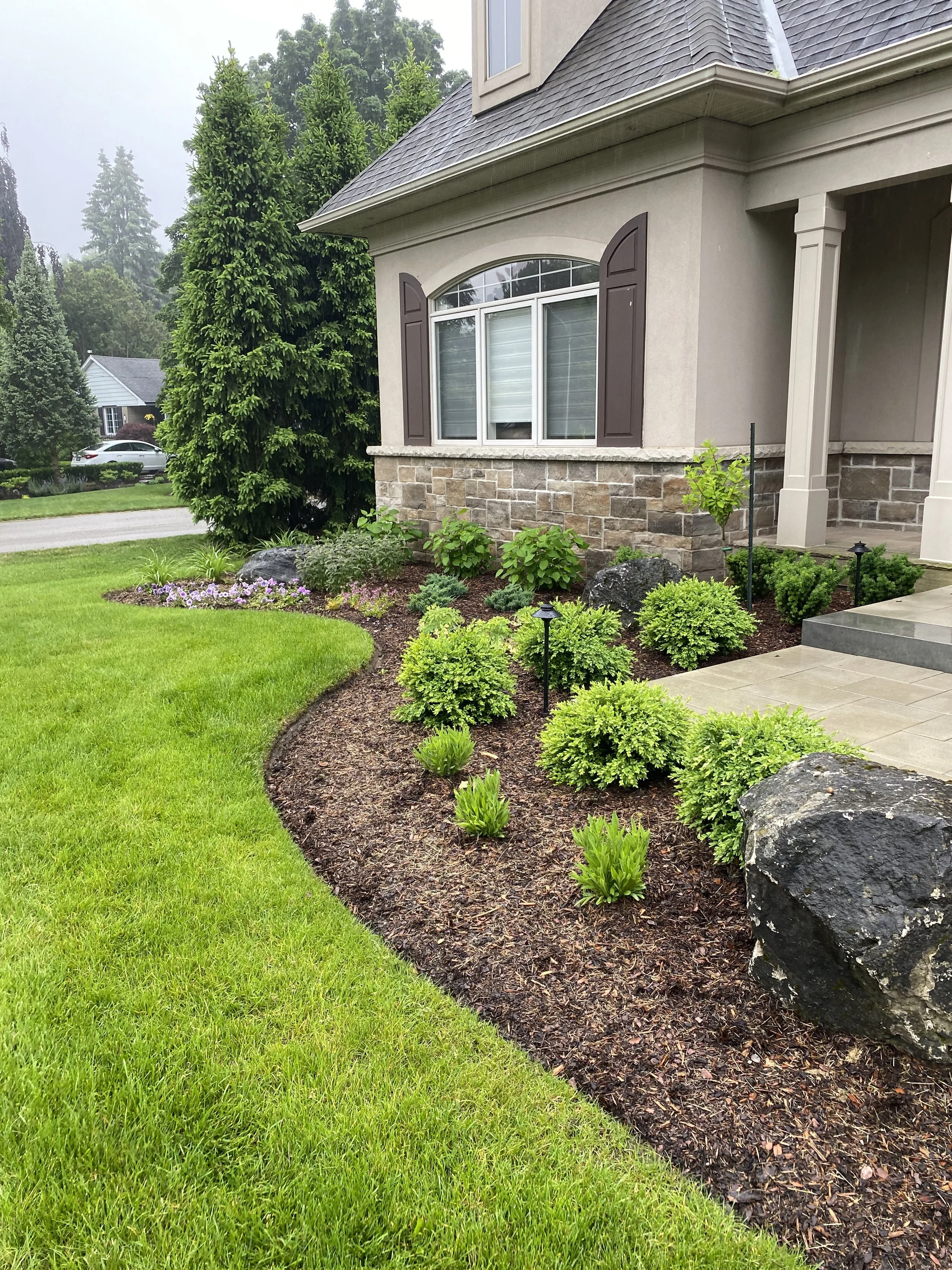 Front yard with a well-maintained garden bed, green shrubs, decorative rocks, and a house with vinyl siding, stone accents, and decorated shutters.