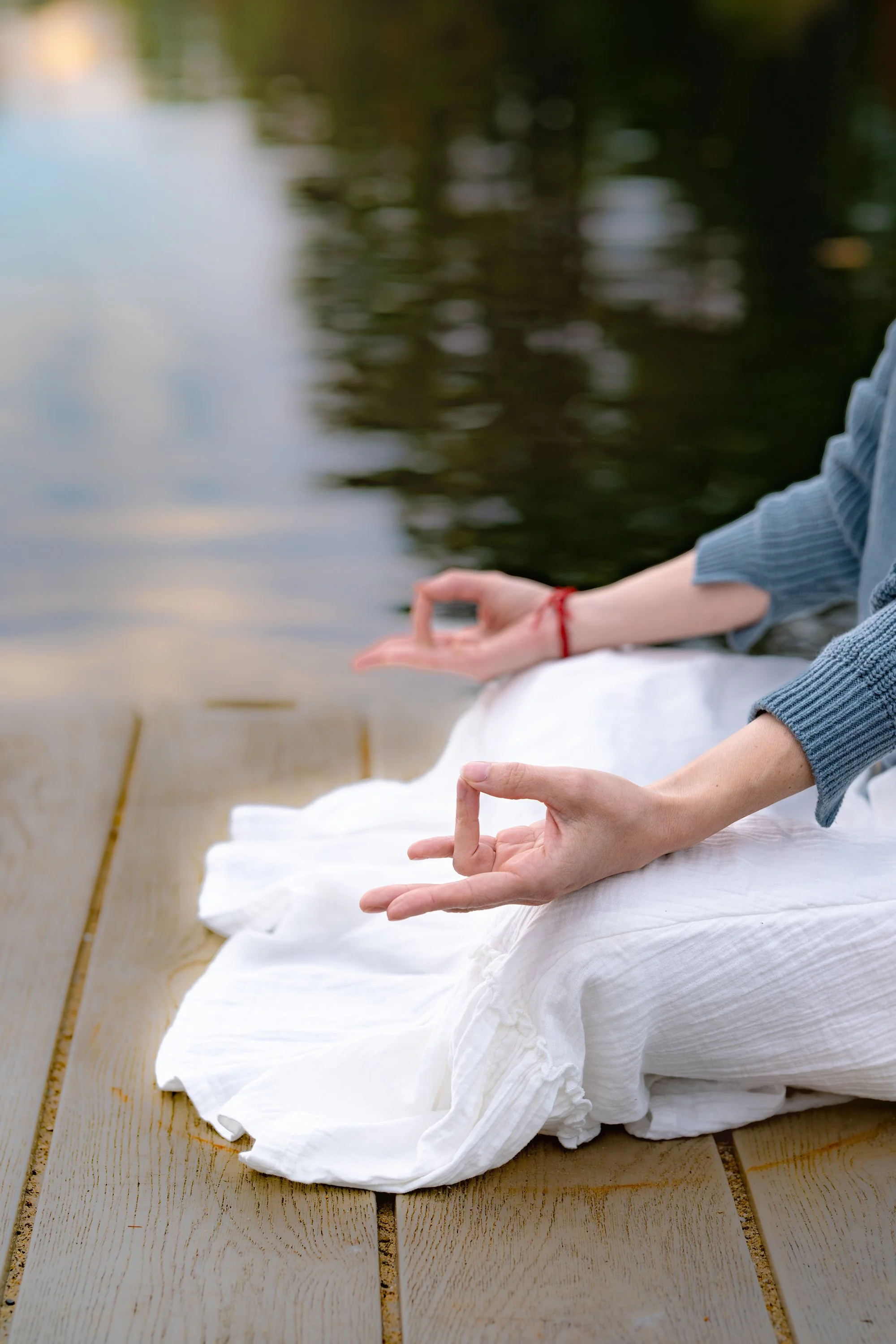 Person sitting cross-legged on a wooden deck near water, meditating with hands in Prthivi Mudra (earth gesture), wearing a blue long-sleeve shirt and white skirt, with a serene outdoor setting.