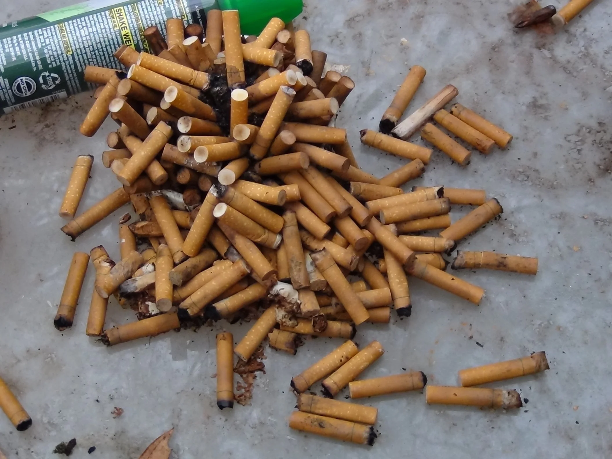A pile of cigarette butts scattered on a concrete surface next to an almost empty cigarette pack.