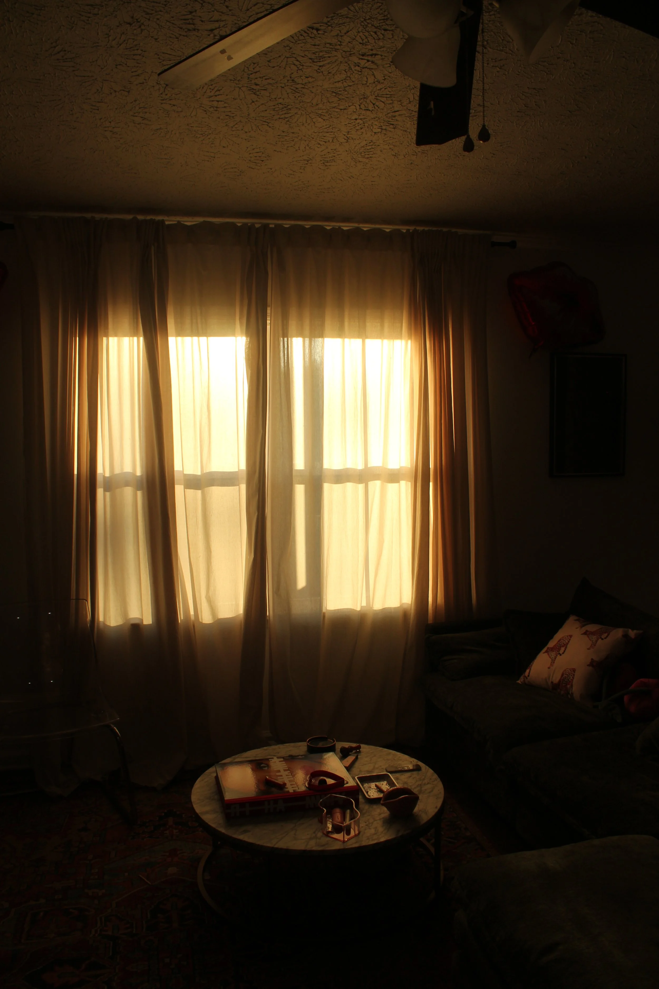 Living room with sunlight shining through beige curtains, a sofa with pillows, a round coffee table with magazines and small decorative items, and a ceiling fan.