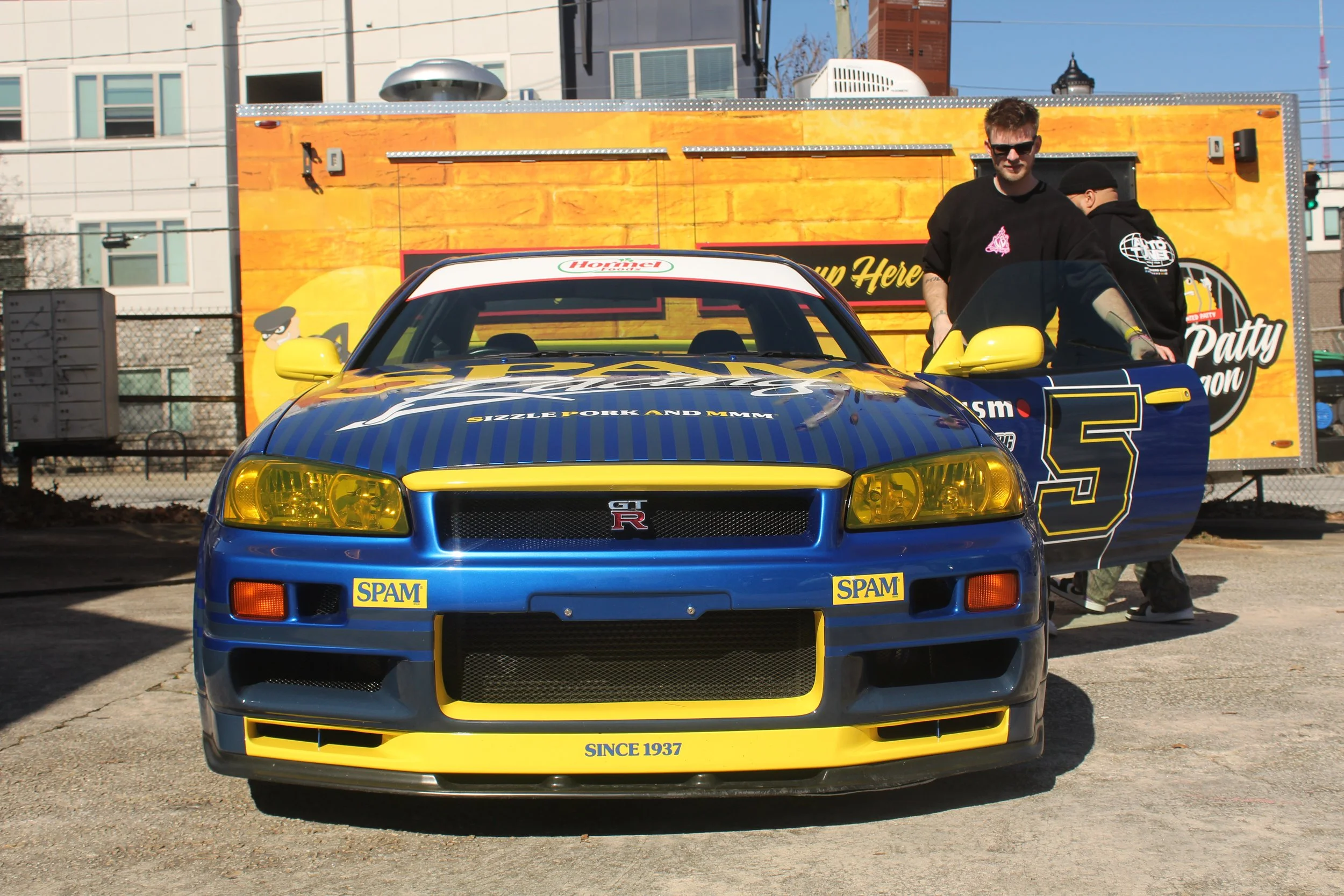 A blue and yellow Nissan GT-R sports car with Spam advertisements on the front, parked outdoors with two men standing nearby and a yellow food truck in the background.
