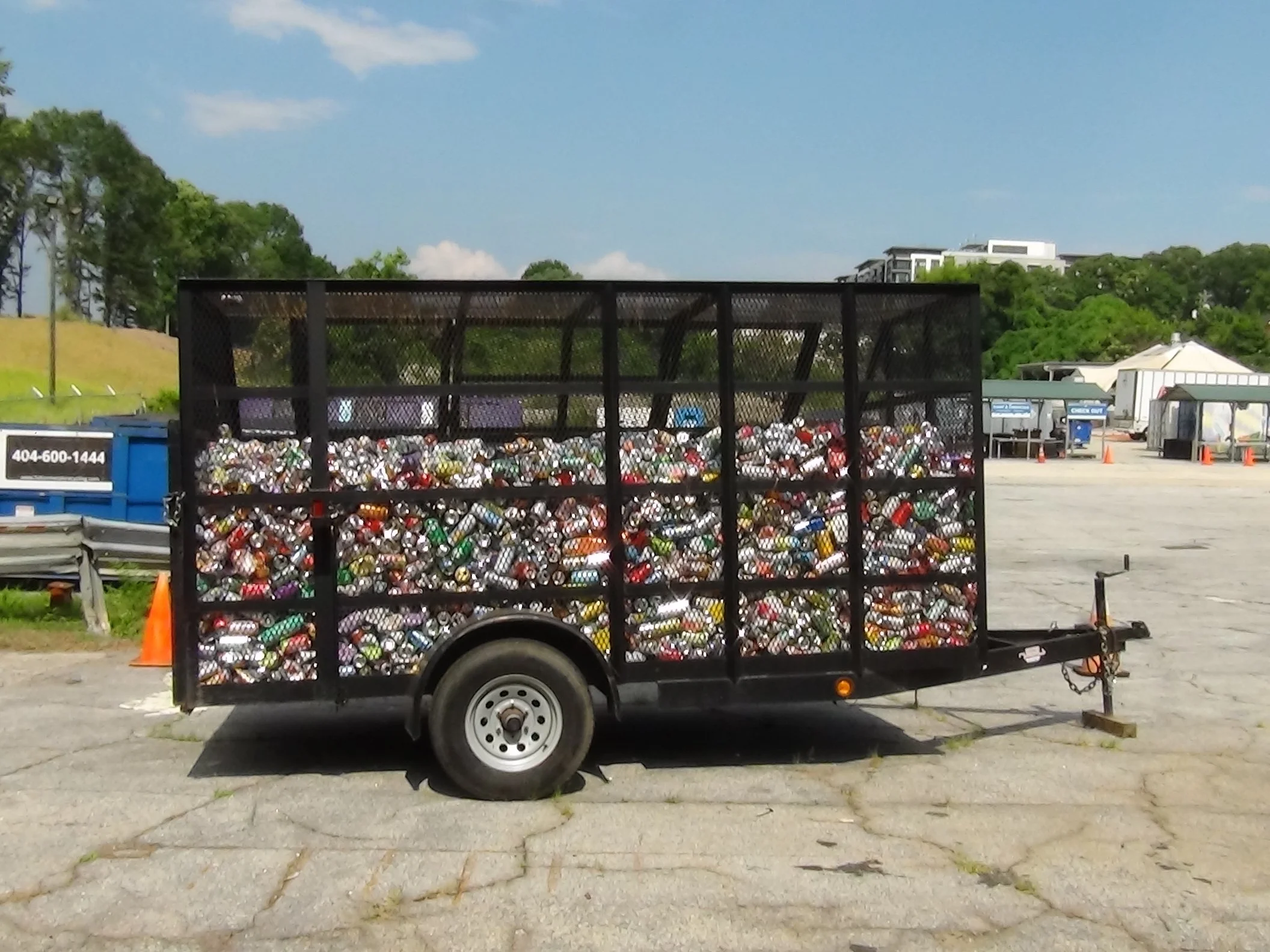 Trailer filled with crushed cans parked in a lot with trees and buildings in the background.