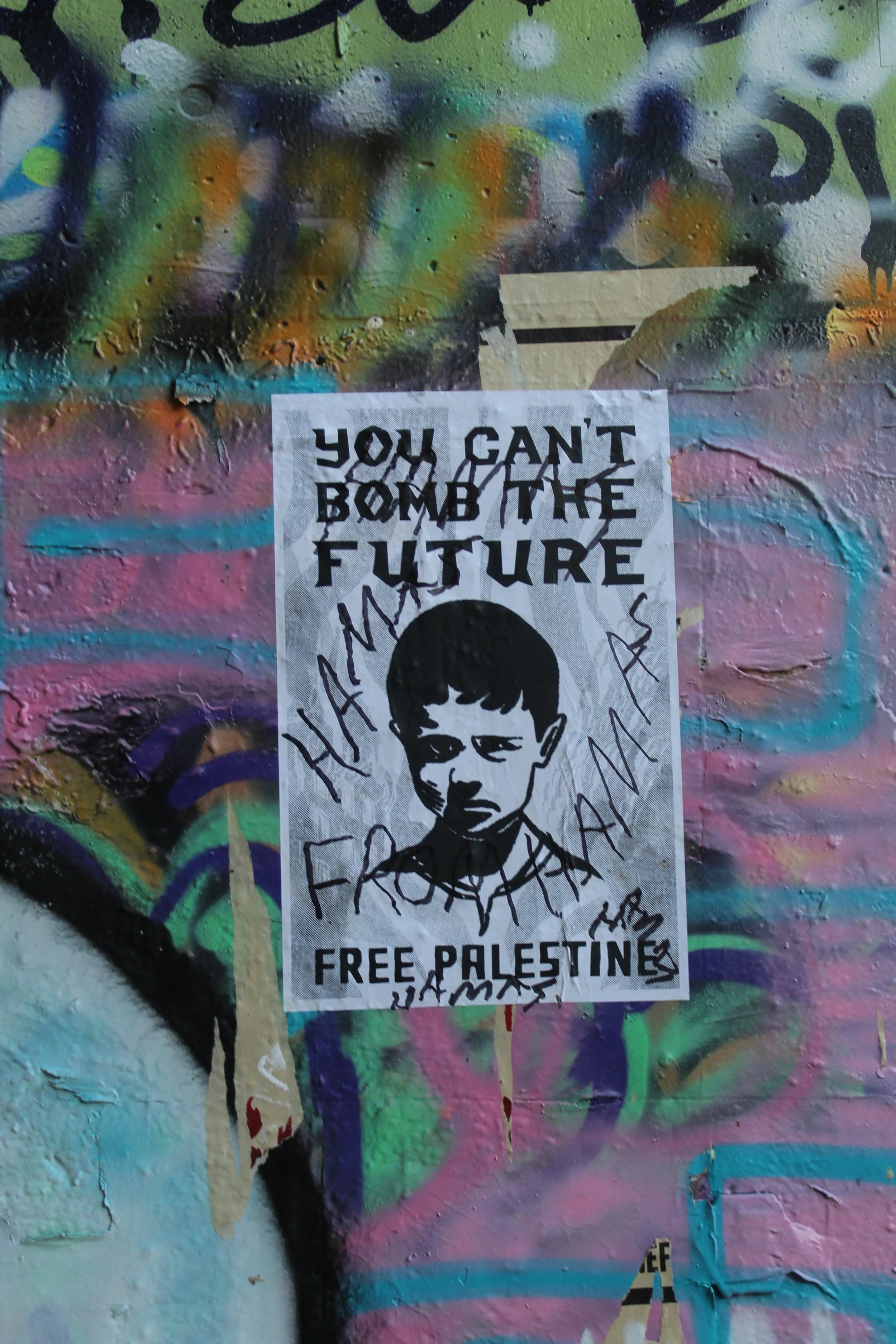 A graffiti-covered wall with a black-and-white poster displaying the phrase 'You can't bomb the future' and a drawing of a young boy. The wall features colorful spray paint and various layered street art.