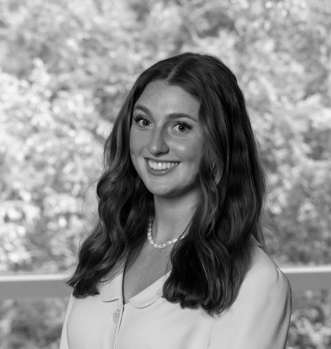 Black and white portrait of a smiling woman with long, wavy hair, wearing a light-colored top, and a pearl necklace, standing outdoors in front of a blurred background of trees.