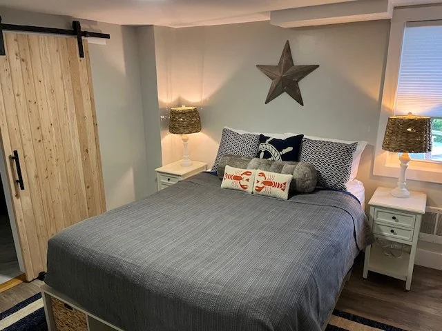 Bedroom with a queen bed, gray bedding, decorative pillows, white nightstands with lamps, a barn door and doorway that leads to a bathroom, a star wall decor, and a window with blinds.