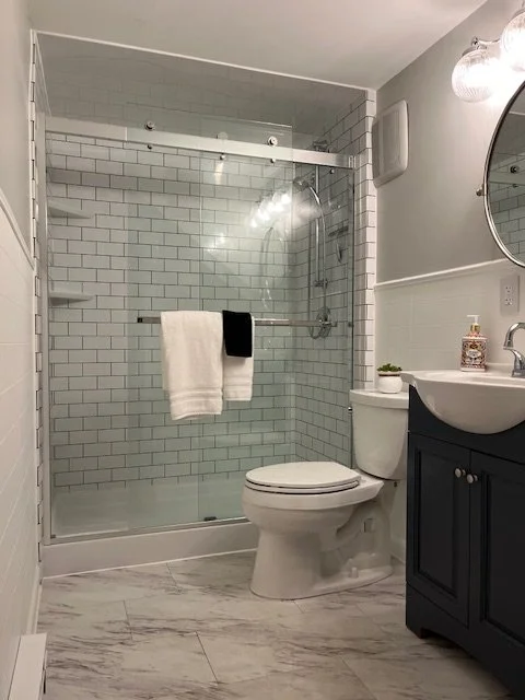 Bathroom with glass shower enclosure, white subway tiles, a toilet, a navy blue vanity, a white sink, and a mirror with light fixtures.