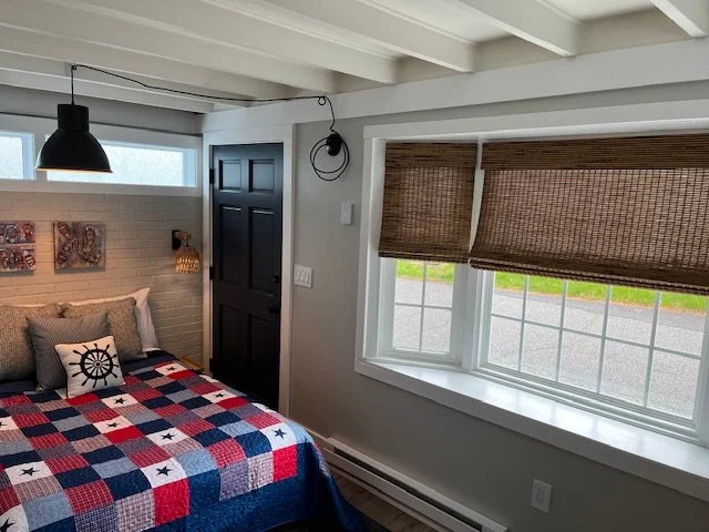 A cozy bedroom corner with a bed covered in a red, white, and blue patchwork quilt, pillows including a decorative one with a ship wheel, a black pendant light hanging from the ceiling, a black door, and a large window with woven bamboo blinds.