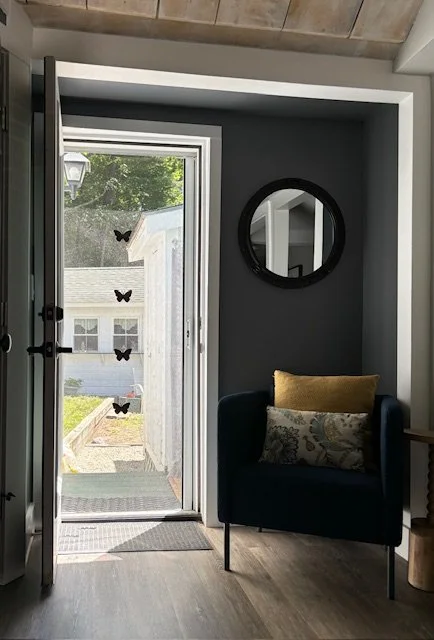 Entryway with open door leading to garden, black armchair with yellow and patterned cushions, round mirror on dark wall, butterfly decorations on window, wooden flooring, and ceiling with exposed beams.