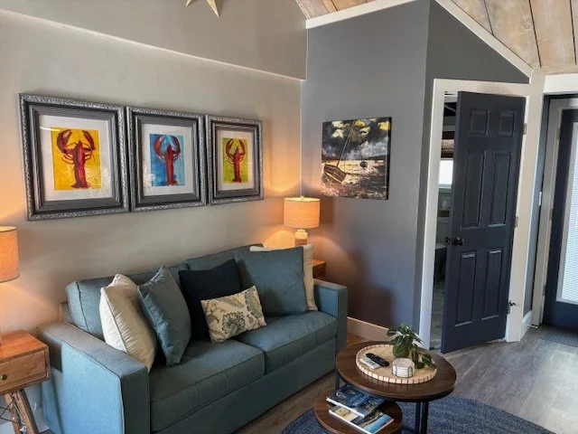 Living room with blue sofa, decorative pillows, three framed dog artwork, two lamps, a wooden side table, a round coffee table with books and a plant, and an open door leading outside.