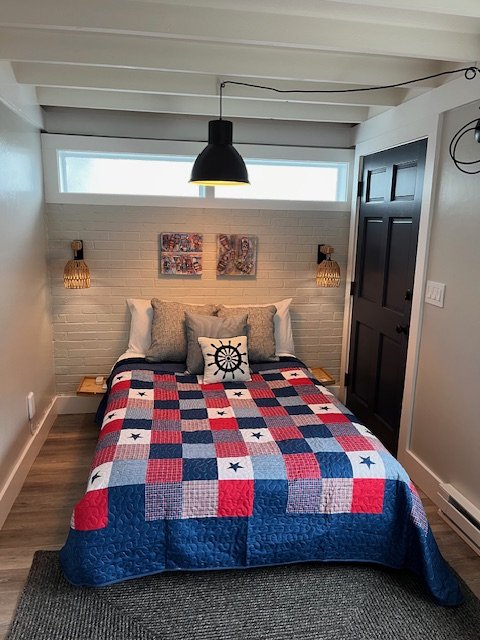 Bedroom with a bed covered in a red, white, and blue quilt, six pillows including one with a ship's wheel, two wall sconces, black ceiling light, and white brick wall with small rectangular windows.
