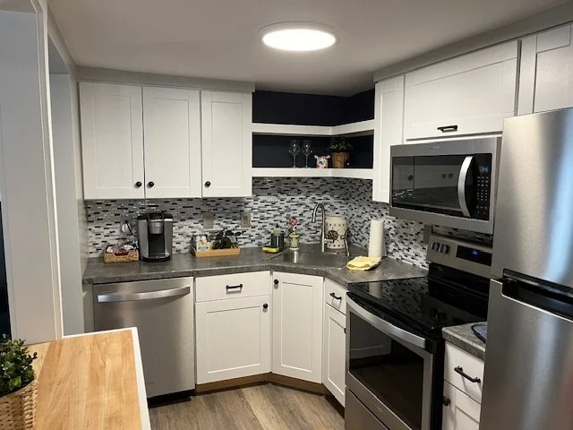 White kitchen with gray countertops, mosaic tile backsplash, stainless steel appliances, and open shelves with glasses and decor.
