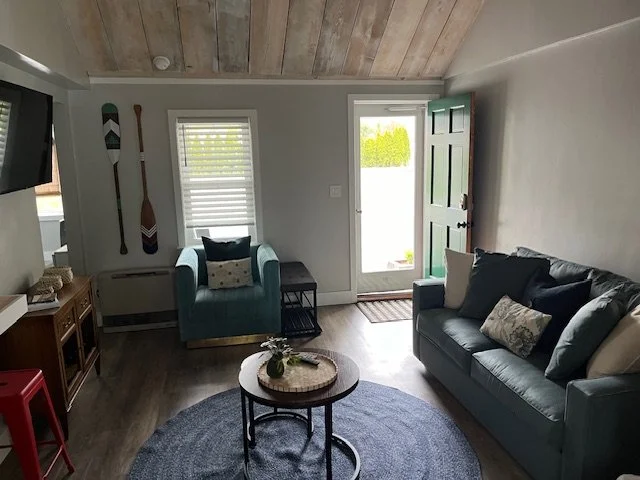 Cozy living room with a teal armchair, and teal sofa with decorative pillows, round coffee table on a blue rug, wall decor with paddles, and a door leading outside, illuminated by natural light.