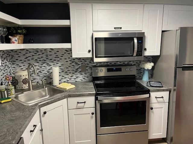 Modern kitchen with white cabinets, stainless steel appliances including microwave, oven, and refrigerator. The countertop has a sink, paper towel hol, and small decorative items. There is a textured tile backsplash with gray, white, and blue tiles.