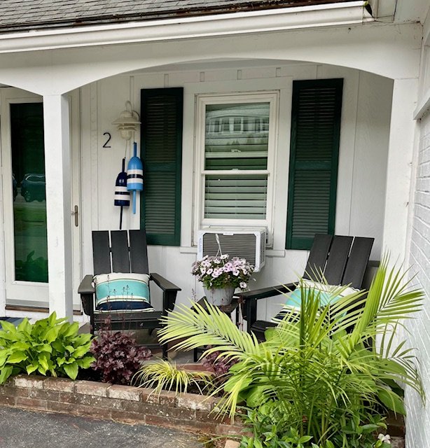 Front porch of a house with two black chairs, a flower pot with pink and white flowers, and green plants. The house has green shutters, a window with blinds, and two decorative buoys hanging on the wall.