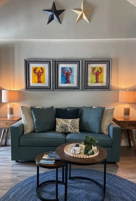 Living room with a teal sofa decorated with pillows, wooden side tables with lamps, framed colorful lobster artwork, star wall decorations, and a round coffee table with plants and books, against a light-colored wall.