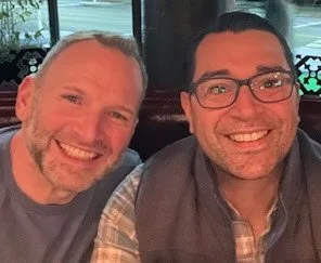 Two men smiling for a photo indoors with a window in the background.