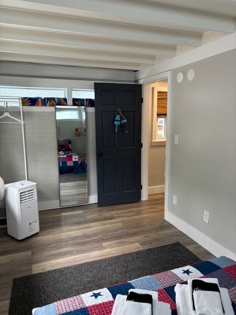 A room with wood flooring, a black door with a coat hanging on it, a window, a wall-mounted heater, and part of a bed with American flag-themed bedding.