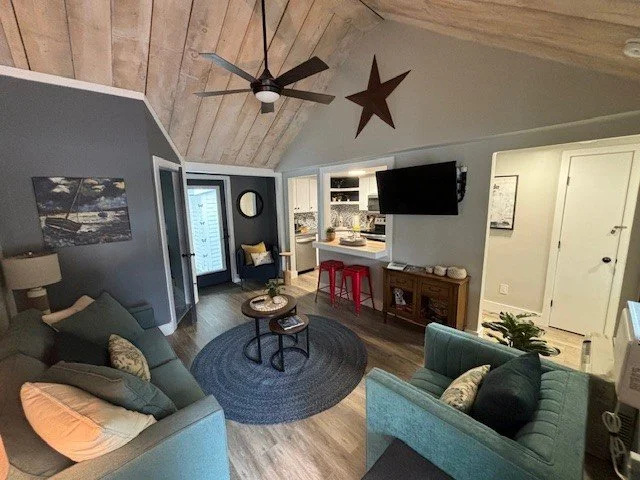 Living room with a vaulted ceiling, wooden paneling, ceiling fan, wall-mounted TV, and a small dining area with a red barstool. There are gray and blue sofas, a round rug, and a small coffee table. The space includes a door with a window, a large star decoration, and a potted plant.