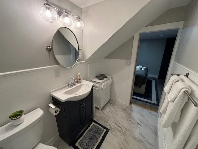 Bathroom with white walls, a sink with a black cabinet, a round mirror, a toilet, a small rug, a towel rack with white towels, and an open door leading to a bedroom.