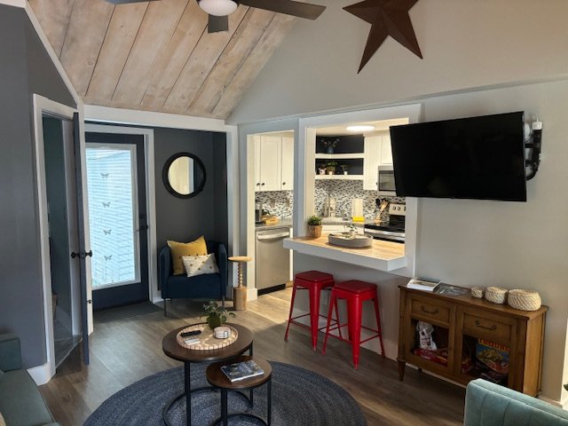 Living room with a vaulted ceiling, TV mounted on the wall, and a view into the kitchen with white cabinetry and a blue backsplash. Decor includes a star-shaped light, a round mirror, a navy armchair with throw pillows, a coffee table with magazines,