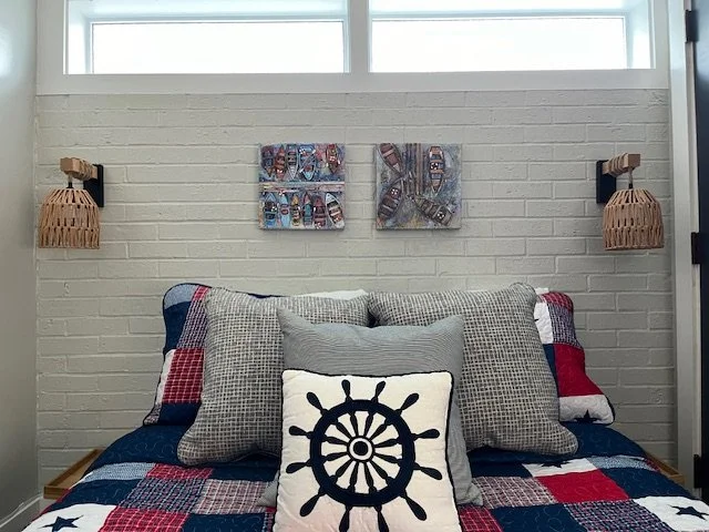 A bedroom wall with two wicker wall sconces and two colorful boat-themed art pieces, a bed with mixed-pattern pillows and a navy, red, and white quilt, and a decorative pillow with a ship's wheel design.