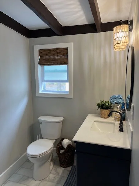 Small bathroom with a white toilet, a window with a brown woven shade, dark wood ceiling beams, a white vanity with a black faucet, a mirror, a basket with toiletries, and blue flowers, illuminated by a wall-mounted wicker sconce.