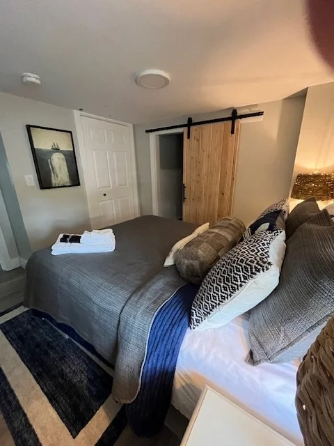 A neatly made bed with multiple pillows, a gray duvet, and a folded towel on top, in a cozy bedroom with a photograph on the wall and a sliding barn door.
