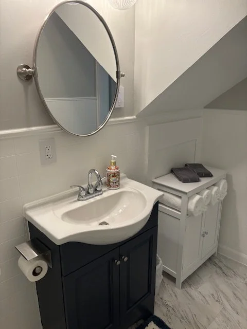Bathroom with a small white sink on a black cabinet, hand soap on the sink, round mirror above, toilet paper holder with toilet paper, white cabinet with folded towels, and black cloth on top, marble floor, and a sloped ceiling.