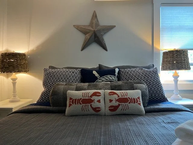 A neatly made bed with a grey bedspread, multiple patterned pillows, and a decorative throw pillow with lobster designs. There are two white bedside tables with matching woven lamps on either side, and a wooden star wall decoration above the bed.
