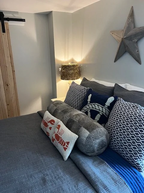 Close-up of a neatly made bed with nautical-themed pillows and a decorative pillow with an anchor, in a cozy bedroom with a star-shaped wall decoration and a textured lamp on the nightstand.