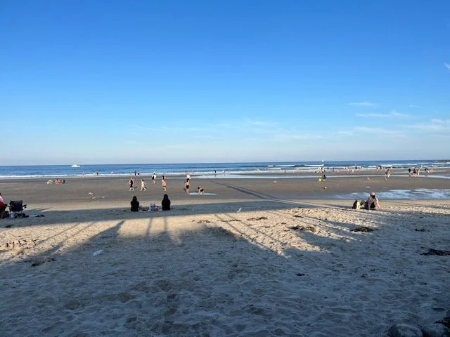 People walking, sitting, and relaxing on a sandy beach with ocean waves in the background, under a clear blue sky.