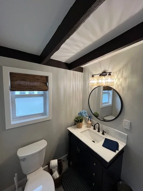 Small bathroom with white walls, a black vanity with a white sink, a round mirror, a window with a brown wicker blind, and decorative blue flowers on the countertop.