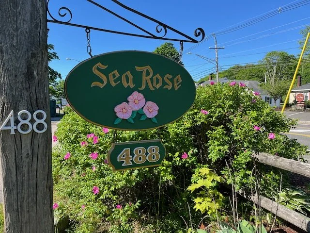 Business sign for Sea Rose with pink flowers, attached to a wooden pole, visible house and road in the background, outdoor scene with greenery and flowering bushes.