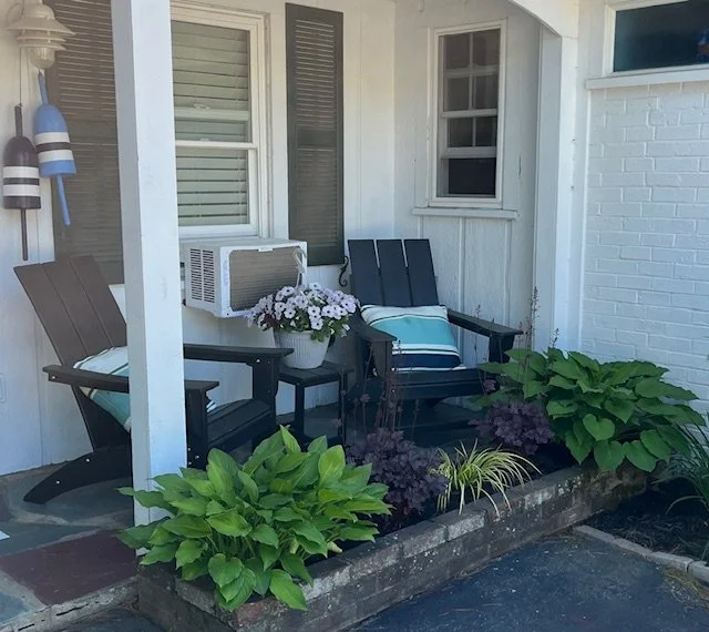 Porch with two chairs, a small table with a flower pot, and garden plants in front.