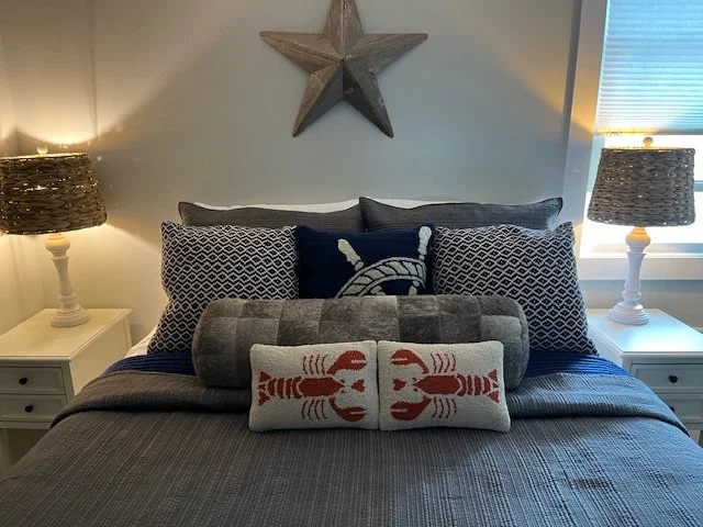 A neatly made bed with navy blue, gray, and white bedding and decorative pillows, including a lobster pillow and a wheel design pillow, flanked by white nightstands with table lamps, in a room with a star decoration on the wall and a window with blinds.