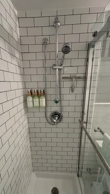 Shower with white subway tiles, a rainfall showerhead, shower controls, a handheld shower wand, a glass door, and three bottles of soap or shampoo.