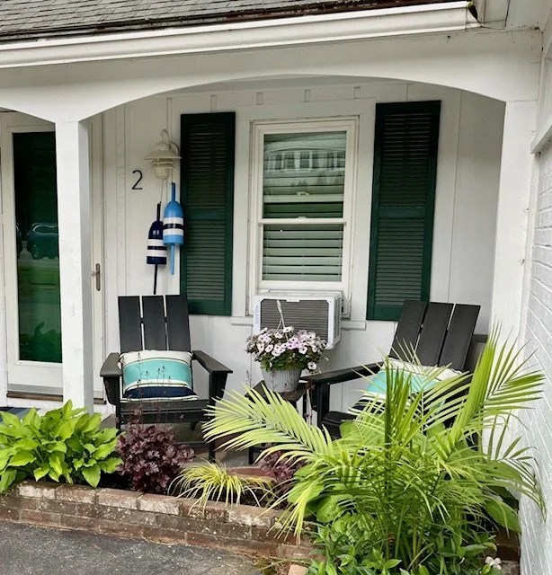 Front porch with two black Adirondack chairs, a small table with a flower pot in the center, green and purple plants in a brick planter, a window with dark green shutters, and decorative blue and white buoys hanging on the wall.