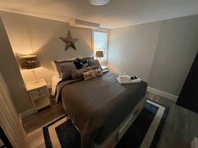 Bedroom with a bed, pillows, pillows with decorative covers, a nightstand, lamps, a star wall decoration, window, and towels laid out on the bed.
