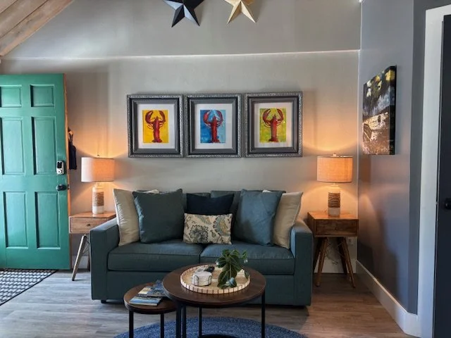 Living room with a teal sofa, colorful lobster artwork framed on the wall, two wooden side tables with lamps, and a round coffee table with a plant and magazines, with star decorations hanging from the ceiling.
