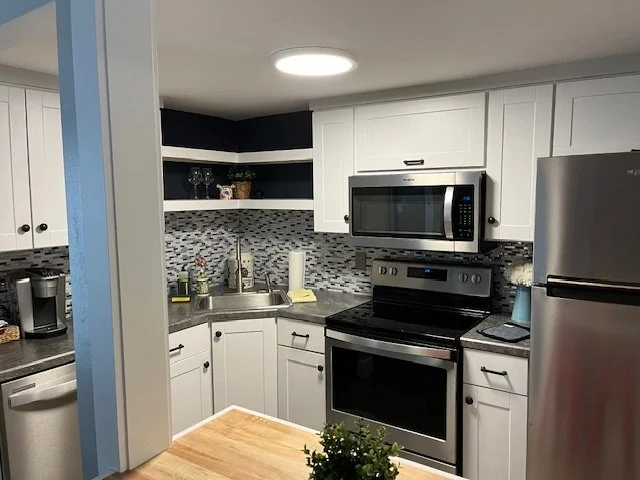 Kitchen with white cabinets, stainless steel microwave and refrigerator, black oven, mosaic tile backsplash, and grey countertop.