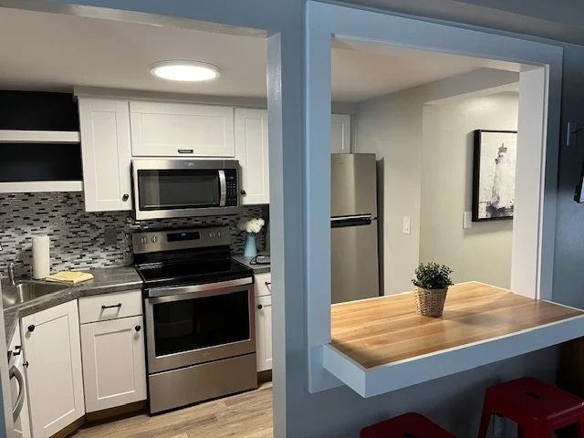 View of a modern kitchen with stainless steel appliances, white cabinets, a mosaic tile backsplash, and a bar-like dining area with stool seating