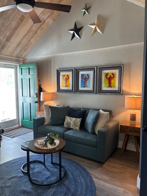 Living room with teal sofa, decorative pillows, wooden side tables with lamps, framed artwork on the wall, star-shaped decorations on the ceiling, a blue circular rug, and a window with a forest green door.