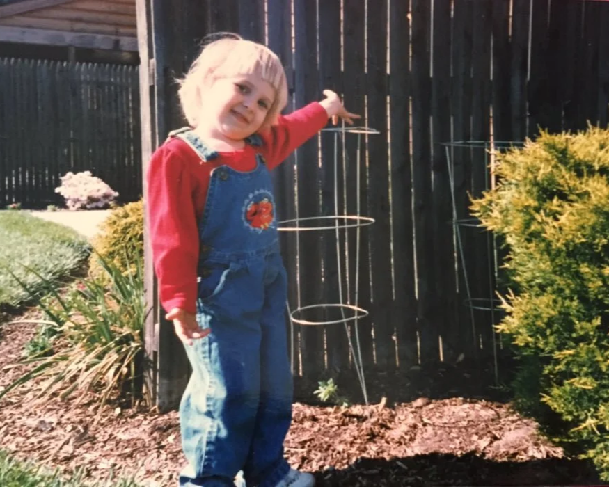 A young girl with blonde hair is standing outdoors, smiling and pointing towards a tomato plant.