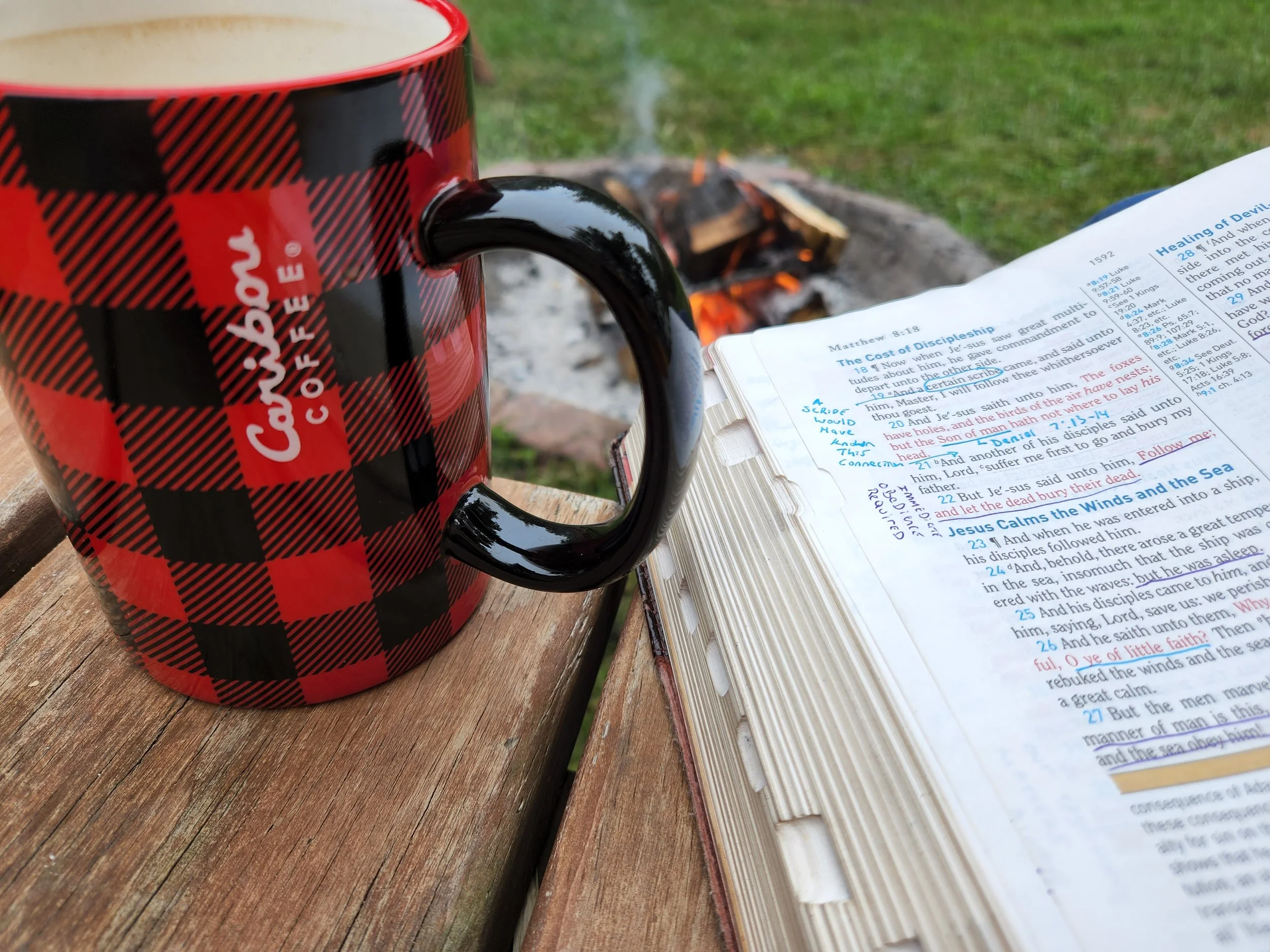 A red and black plaid coffee mug with the word 'Caboose' on it, placed on a wooden surface next to an open Bible, with a campfire burning in the background on a grassy area.