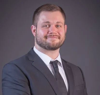 Professional portrait of a man in a dark suit, white shirt, and tie, with short hair and beard, smiling, against a gray background.