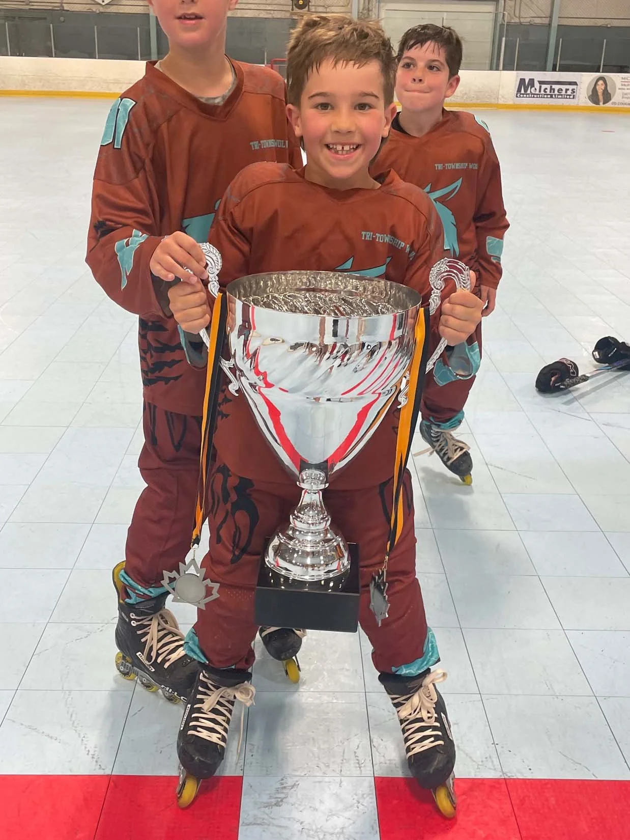 Three children in hockey gear on ice rink, one boy in front holding a large silver trophy with medals, smiling.