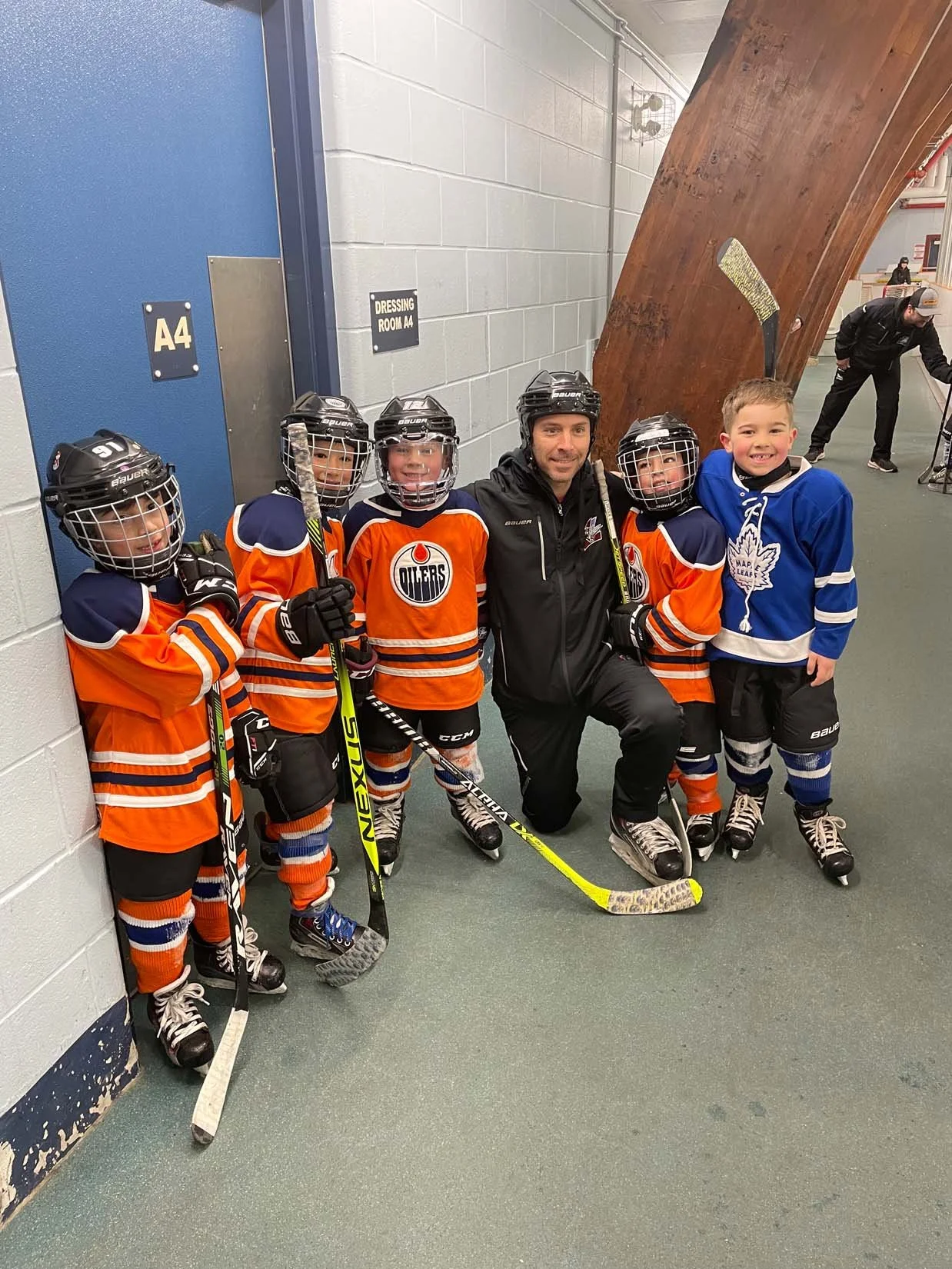 Hockey coach and young players in orange and blue jerseys posing against the wall in an indoor hockey rink.