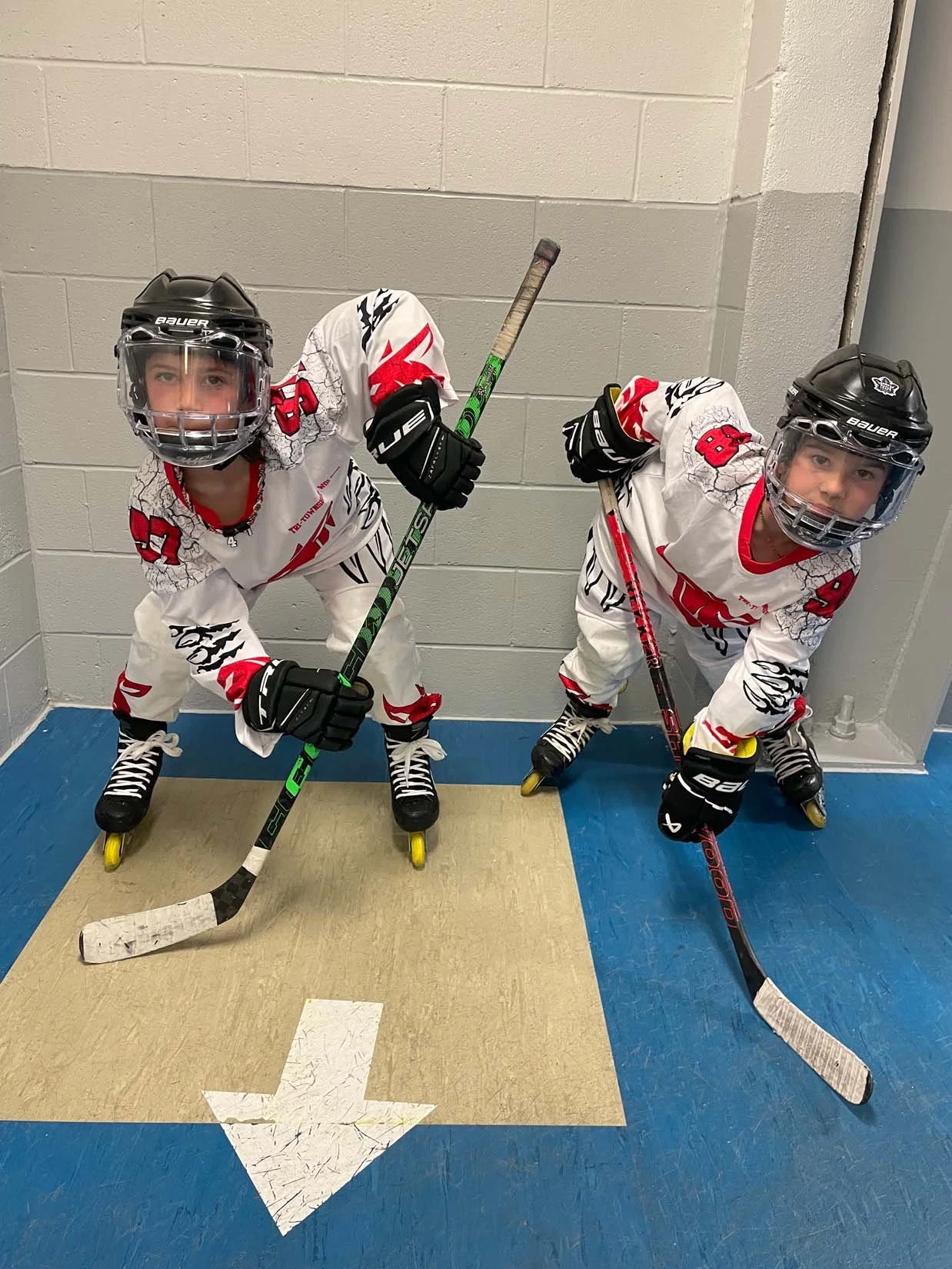 Two young boys in hockey gear and uniforms, wearing helmets, standing on rollerblades, holding hockey sticks, and posing for a photo in an indoor setting with a painted arrow on the floor pointing downward.