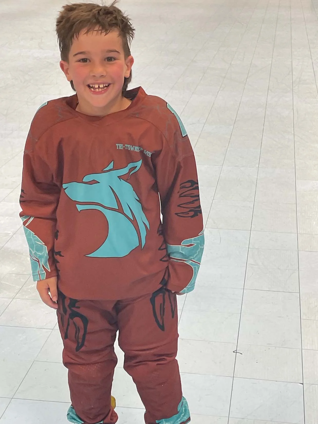 A young boy smiling while standing on a tiled floor, wearing a brown and teal sports jersey and matching pants.